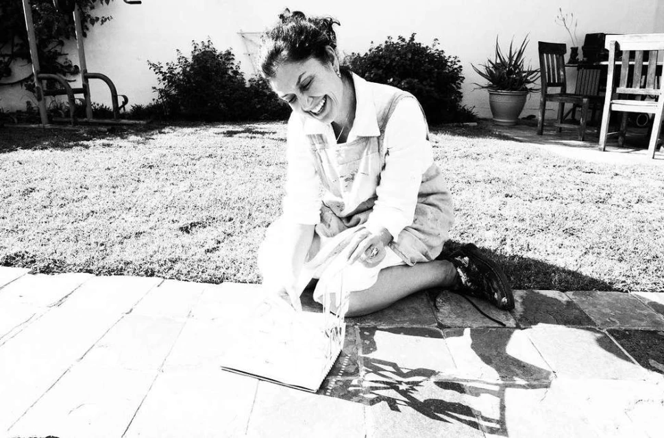 Woman smiles outdoors in black and white, kneeling on stone by a book. Grass and greenery fill the background.