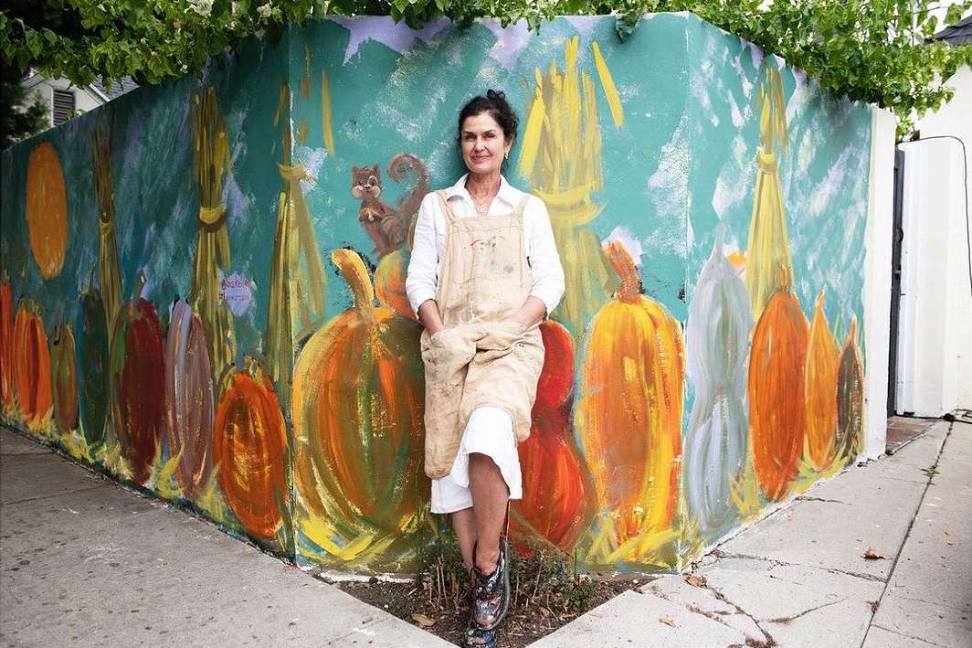 Adult woman stands in front of a colorful outdoor mural with pumpkins, corn stalks, a squirrel, and a light blue background.
