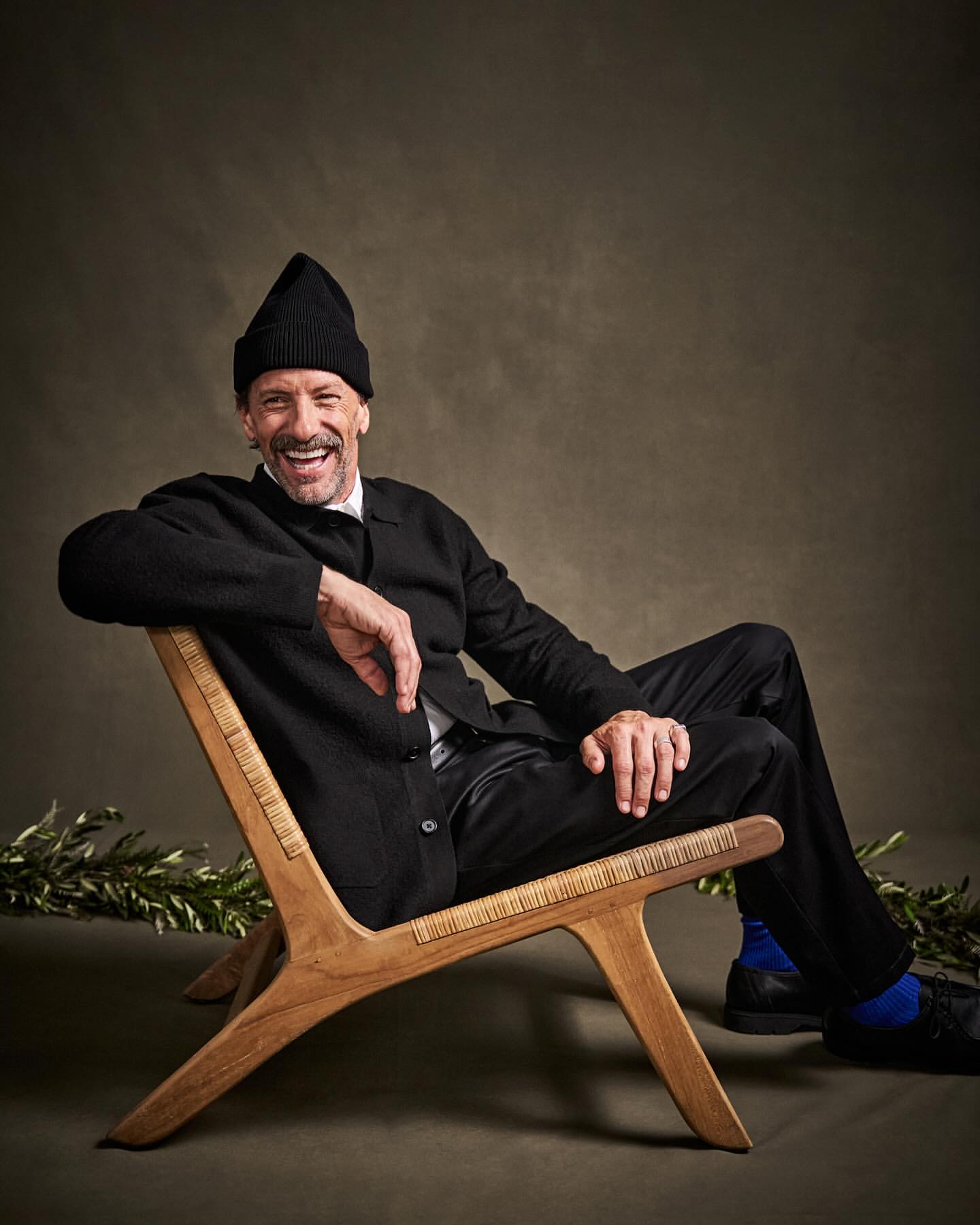 Smiling mature man in a black beanie and cardigan sitting in a wood chair in a studio setting with olive branch on the ground.