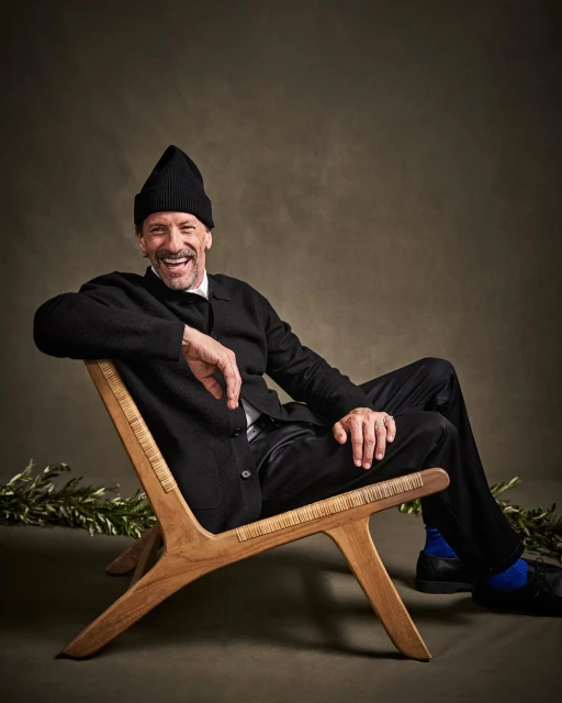 Smiling mature man in a black beanie and cardigan sitting in a wood chair in a studio setting with olive branch on the ground.