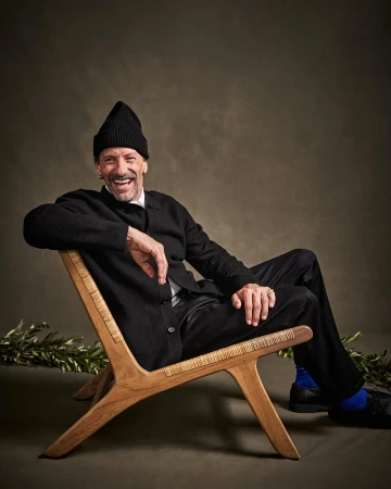 Smiling mature man in a black beanie and cardigan sitting in a wood chair in a studio setting with olive branch on the ground.