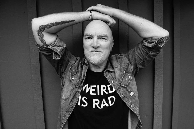 Portrait of a bald man with a beard and tattoos wearing a graphic t-shirt and denim shirt, hands clasped behind his head.