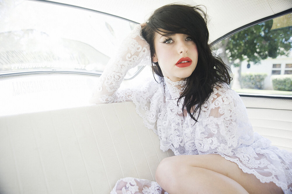 Brunette woman with red lipstick and white lace dress sitting in the backseat of a car with a residential view through the window.