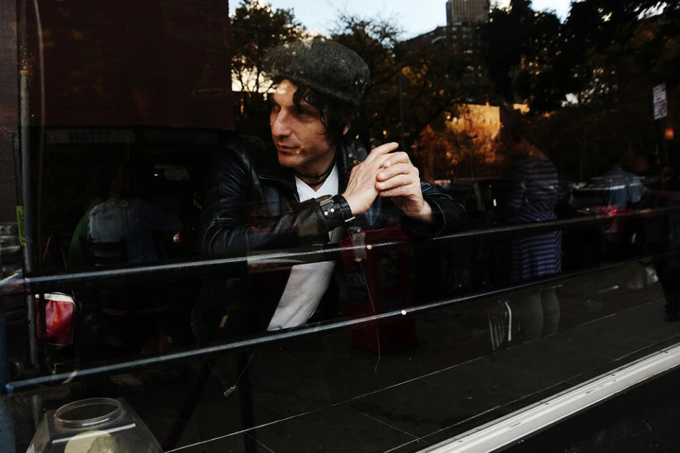An adult male in a black leather jacket and cap, visible through a window with reflections of an urban environment and building.