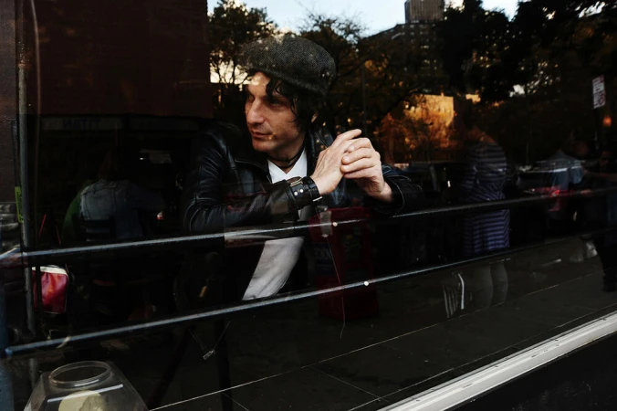 An adult male in a black leather jacket and cap, visible through a window with reflections of an urban environment and building.