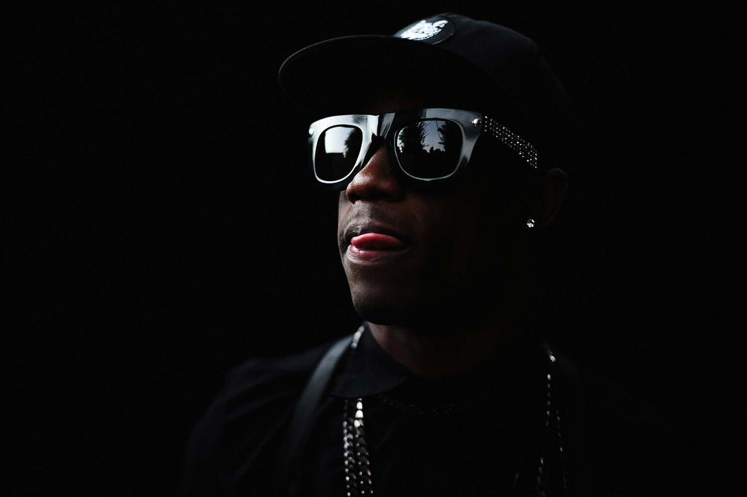 Portrait of a Black adult male wearing sunglasses and a baseball cap against a dark background, sticking his tongue out slightly.