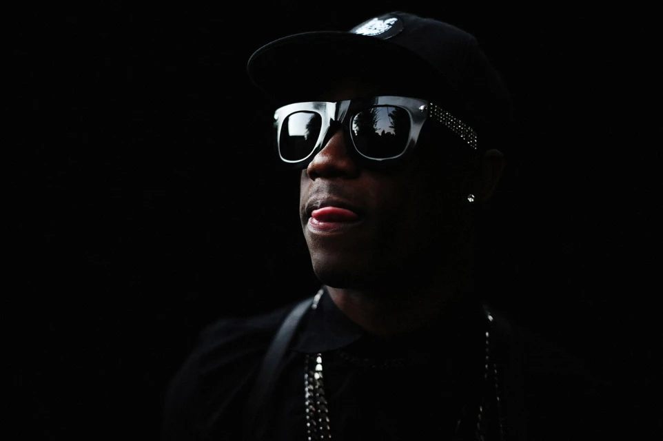 Portrait of a Black adult male wearing sunglasses and a baseball cap against a dark background, sticking his tongue out slightly.