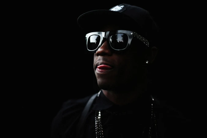 Portrait of a Black adult male wearing sunglasses and a baseball cap against a dark background, sticking his tongue out slightly.