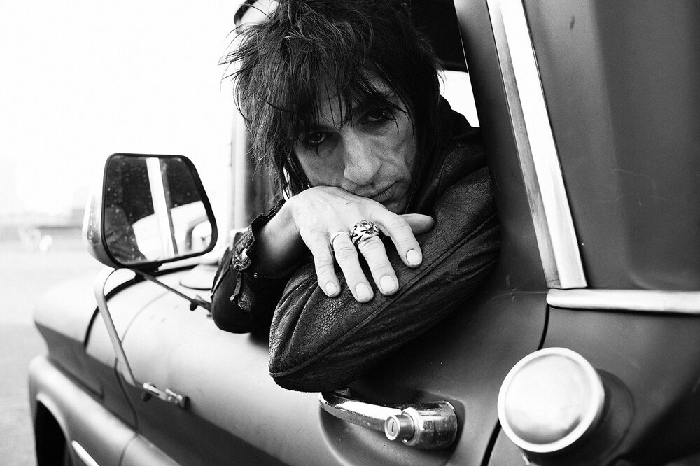 Black and white portrait of Ronnie Wood leaning out of a truck window with dark hair and rings on his fingers by Jeff Forney