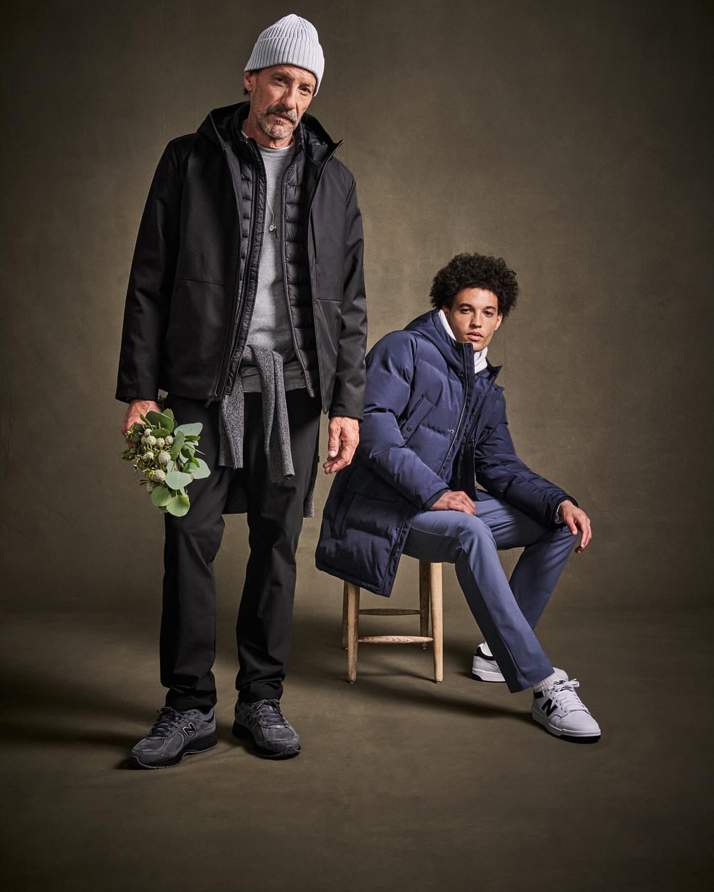 Two adult males, one standing holding flowers wearing a black jacket and beanie, the other seated in a blue puffer jacket.