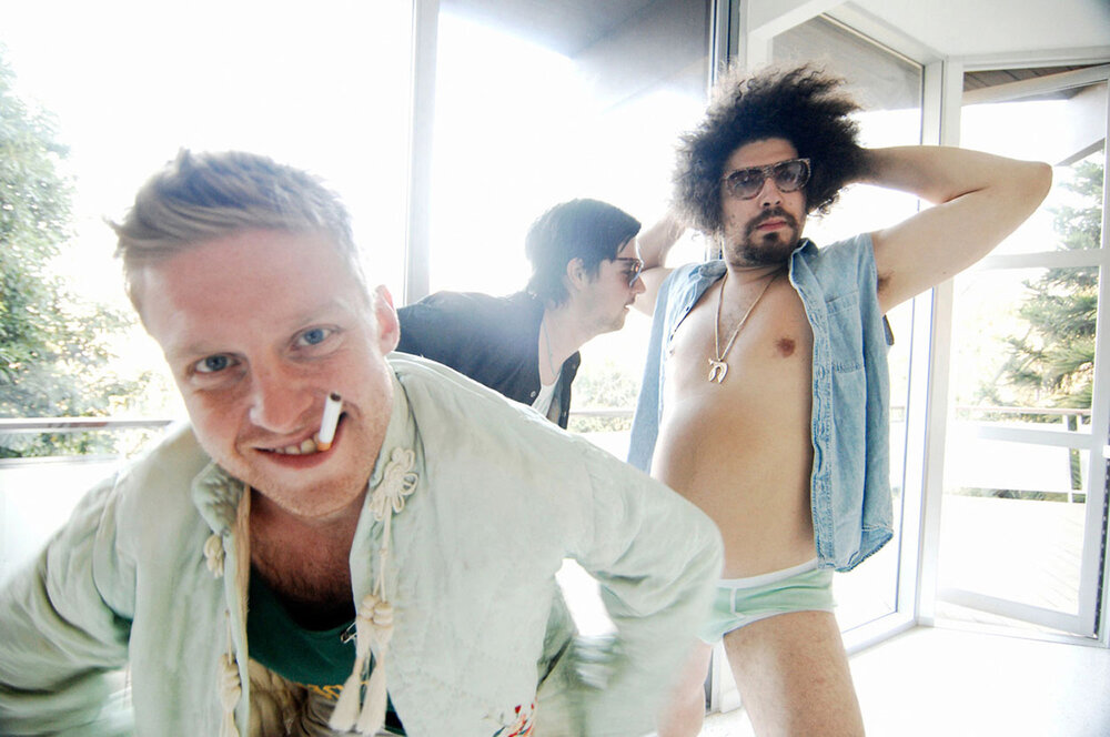 Three adult males pose indoors near a window, wearing casual clothing and accessories, with one man holding a cigarette in his mouth.