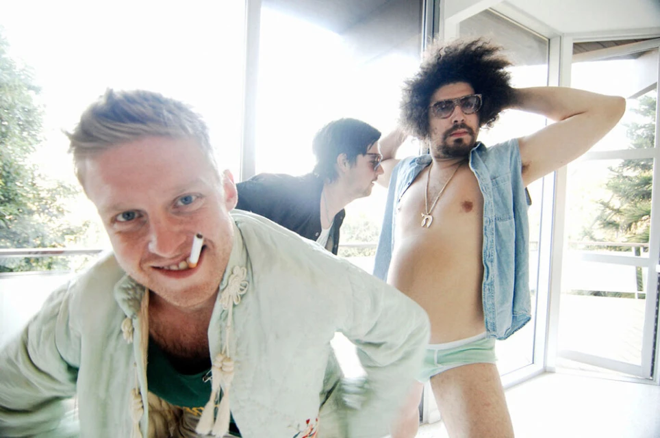 Three adult males pose indoors near a window, wearing casual clothing and accessories, with one man holding a cigarette in his mouth.