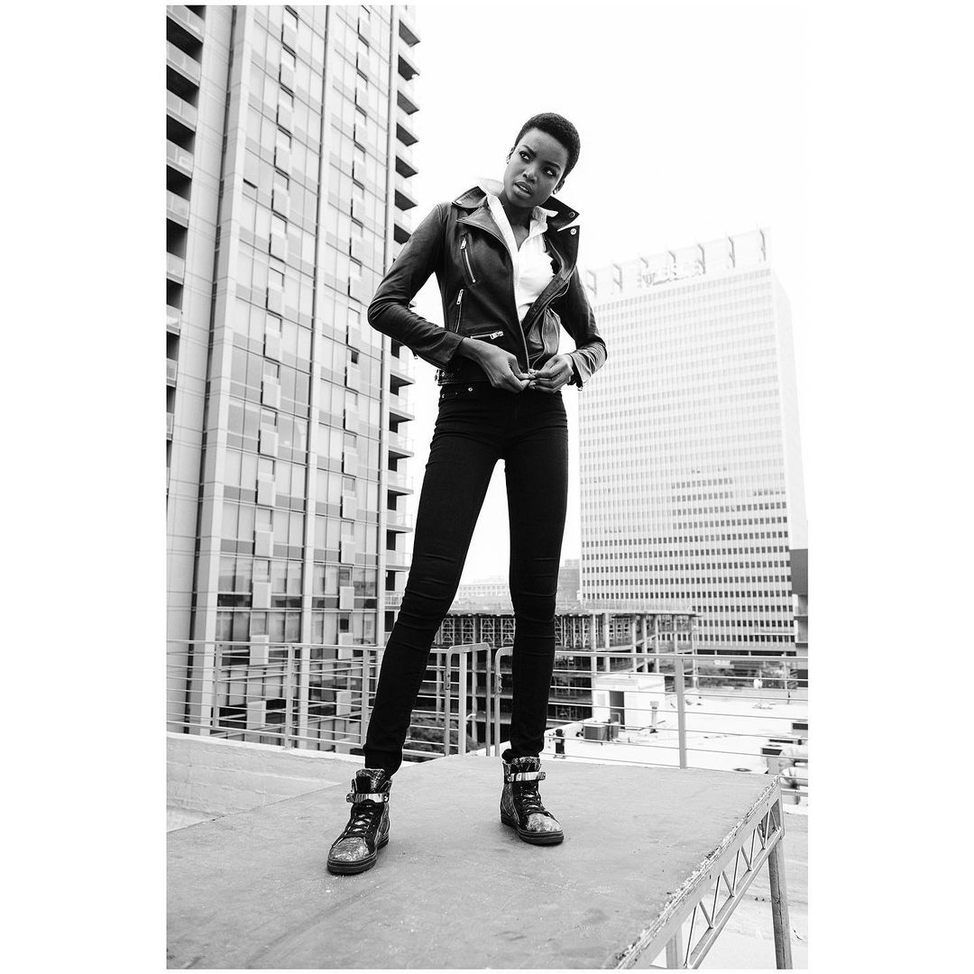 Black and white outdoor fashion portrait of a young woman in a leather jacket, jeans, and high-top sneakers, posing against a city building.