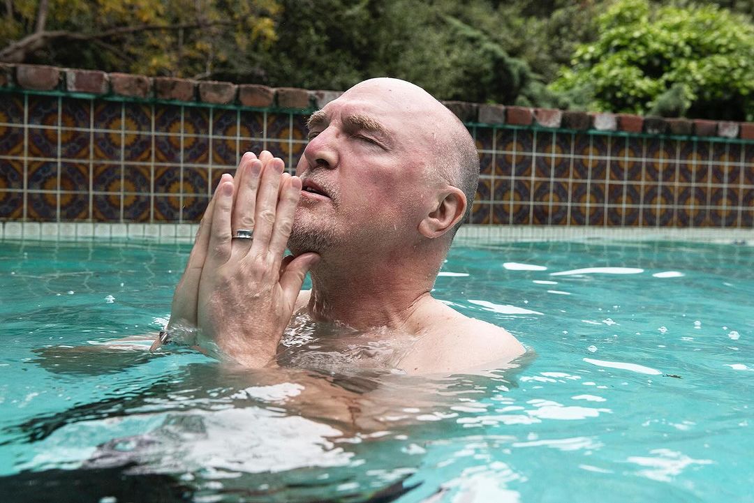 Man in a swimming pool, eyes closed with hands together, creating a serene image with a colorful tiled wall in the background.