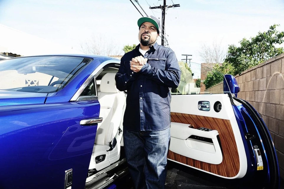 Ice Cube stands next to his royal blue luxury car, showcasing its open door and a modern, upscale design in an outdoor setting.