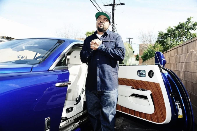 Ice Cube stands next to his royal blue luxury car, showcasing its open door and a modern, upscale design in an outdoor setting.