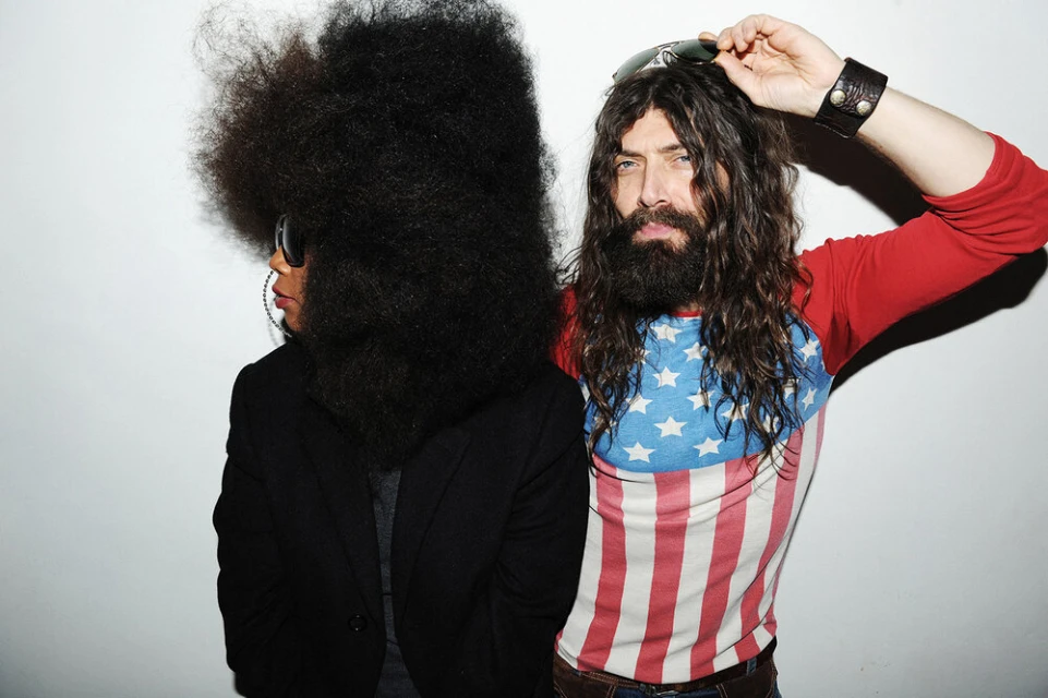 A man and woman pose in front of a white wall. The man wears an American flag shirt and the woman has a very large afro.