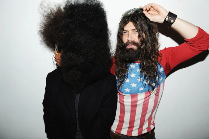 A man and woman pose in front of a white wall. The man wears an American flag shirt and the woman has a very large afro.