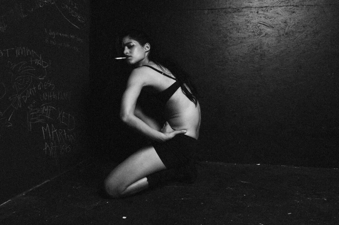 Monochrome portrait of a woman kneeling in a dark corner, holding a cigarette, showcasing a black bra top and shorts.