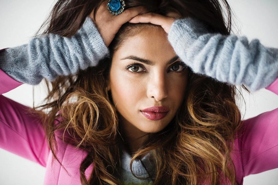 Close-up portrait of a woman with long brown hair, wearing a pink top and blue fingerless gloves, hands on her head, and a blue ring.