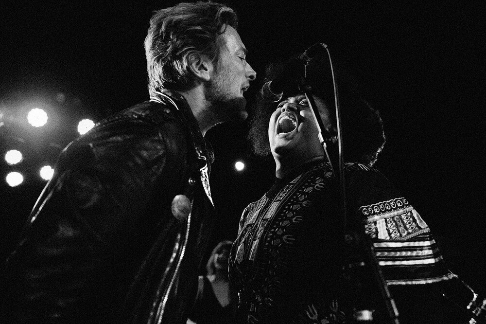 Monochromatic concert photo of two singers: one man with a leather jacket and a woman in patterned top, singing with dynamic expressions.