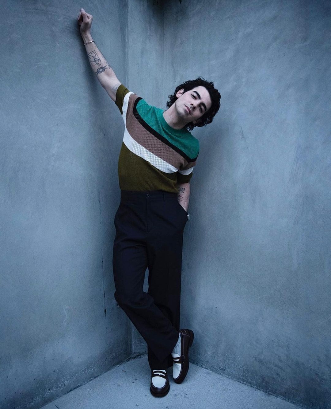 Portrait of a young adult male leaning against a grey concrete wall wearing a striped short sleeve t-shirt, black pants, and loafers.