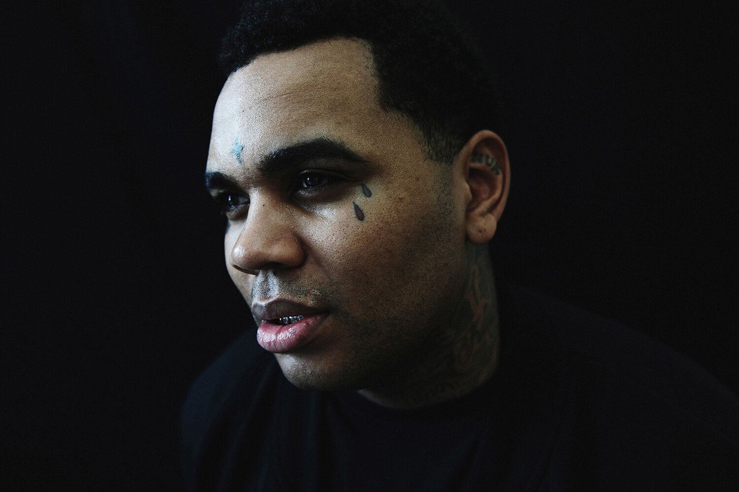 Close-up studio shot of an adult Black man with facial tattoos looking off-camera. He has short dark hair and wears a black top.