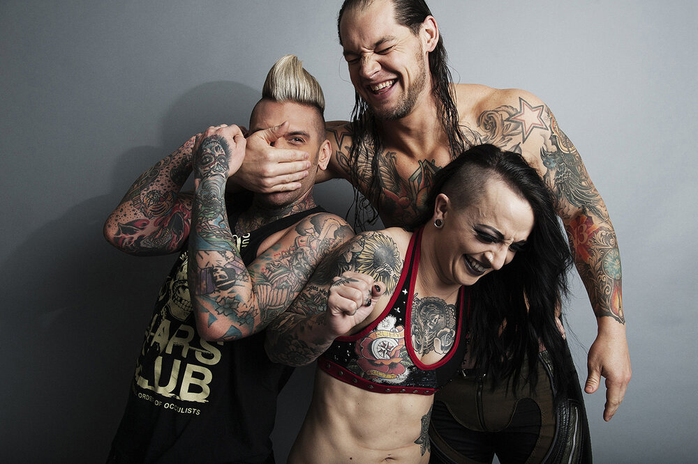 Three tattooed people laughing: one with a mohawk covering his mouth, one bare-chested, and one with a half-shaved head.