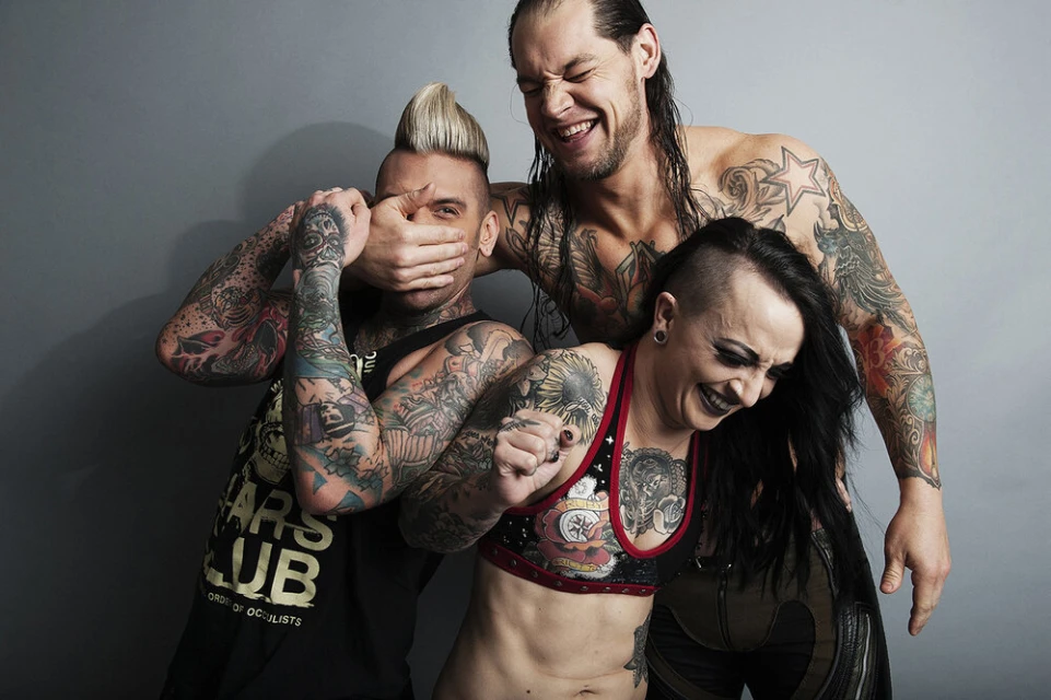 Three tattooed people laughing: one with a mohawk covering his mouth, one bare-chested, and one with a half-shaved head.
