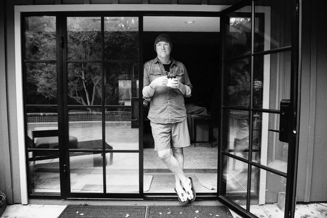 Man standing in open glass sliding doors holding a mug, wearing a beanie, button-up shirt, shorts, and flip-flops.