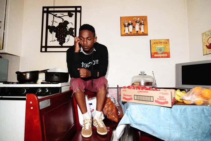 Adult male sitting on a bench in a kitchen wearing a hoodie, shorts, sneakers, and socks, talking on a cell phone, in an indoor setting.