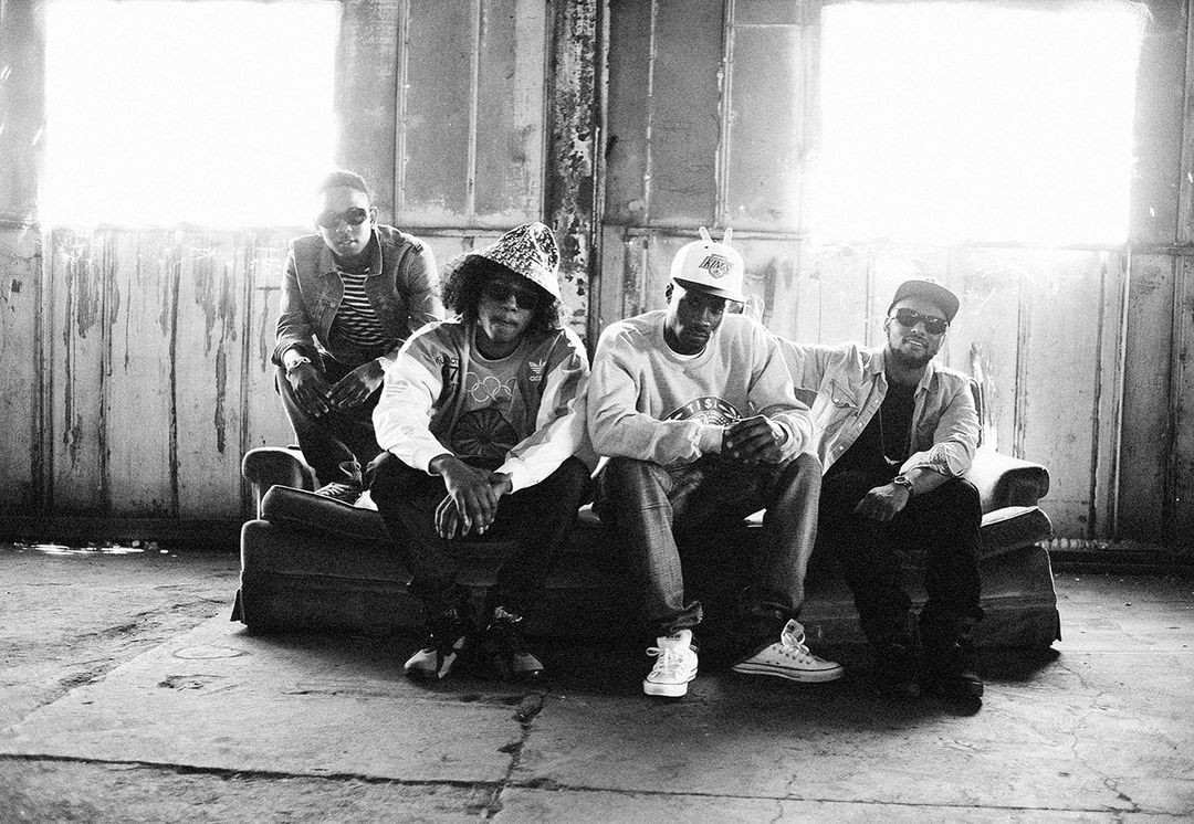 Black and white portrait of four hip-hop artists sitting on a couch in an industrial setting, featuring vintage fashion and accessories.