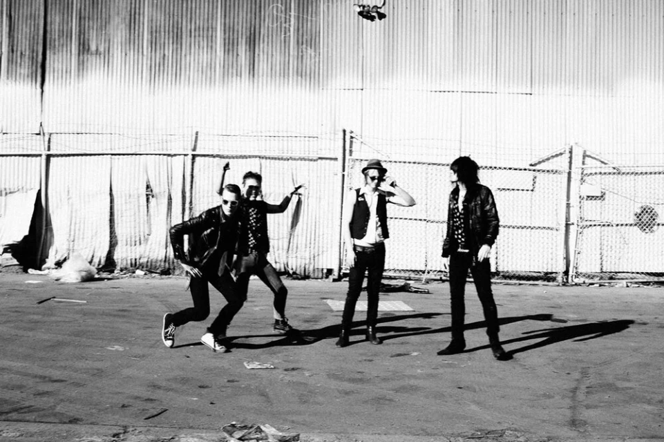 Four people in black jackets pose in an outdoor urban setting with a corrugated metal fence on a sunny day in black and white.