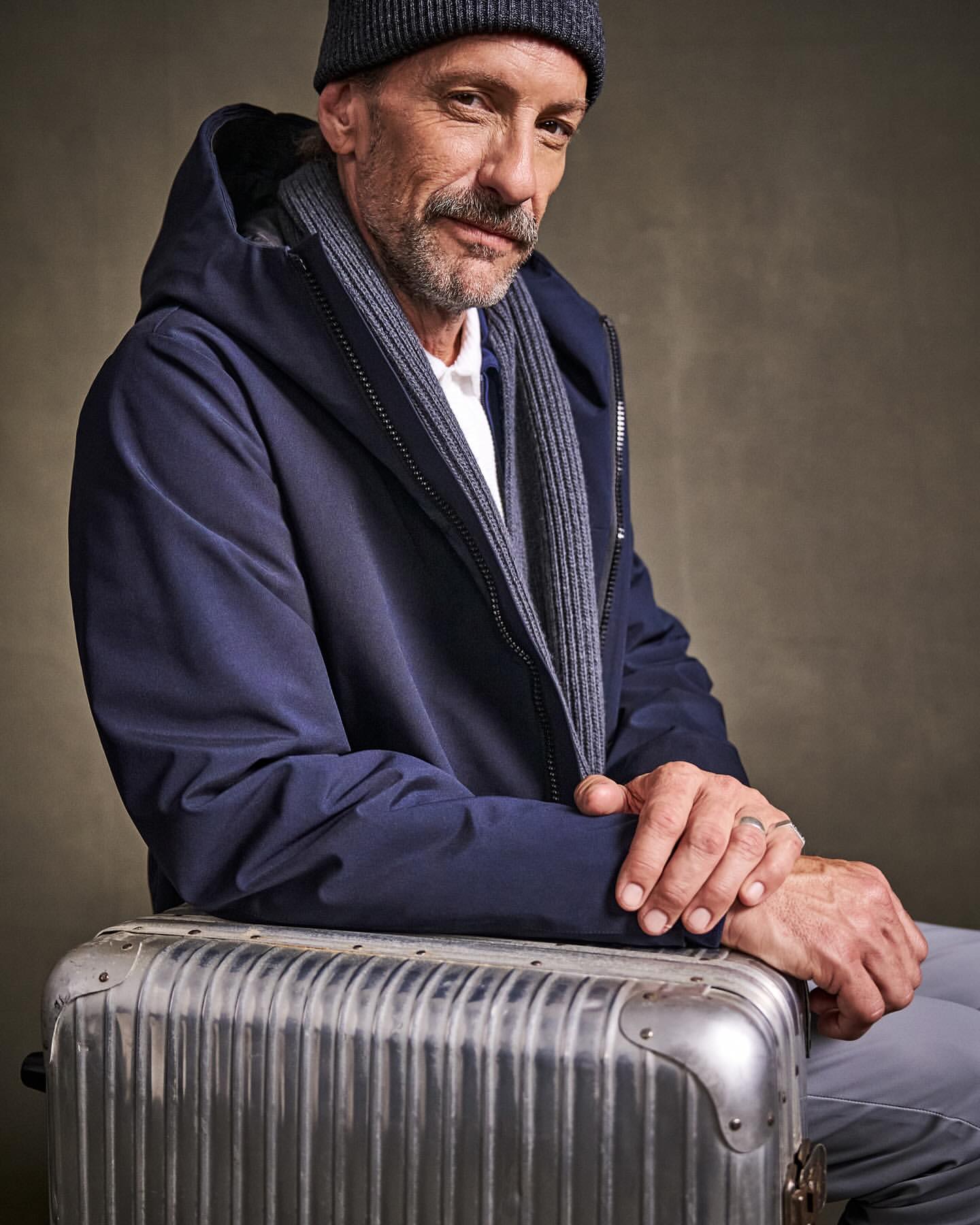 Portrait of a mature man with a mustache, wearing a dark jacket and beanie, seated on a silver metal suitcase.