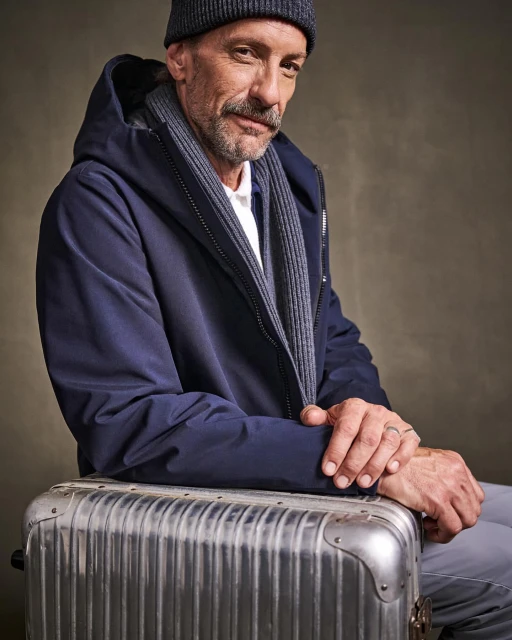 Portrait of a mature man with a mustache, wearing a dark jacket and beanie, seated on a silver metal suitcase.