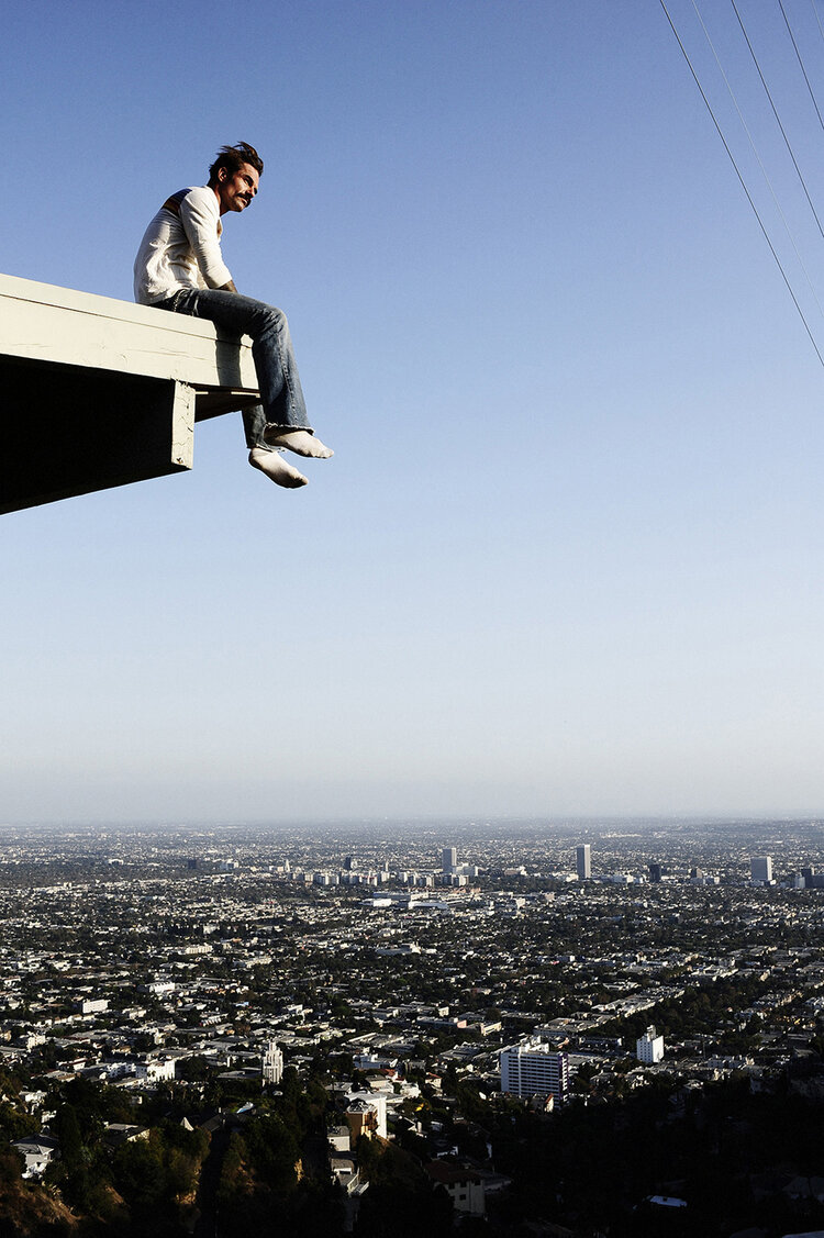 Man sitting on a rooftop edge overlooking a vast sprawling city skyline under a clear blue sky
