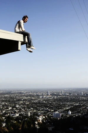 Man sitting on a rooftop edge overlooking a vast sprawling city skyline under a clear blue sky