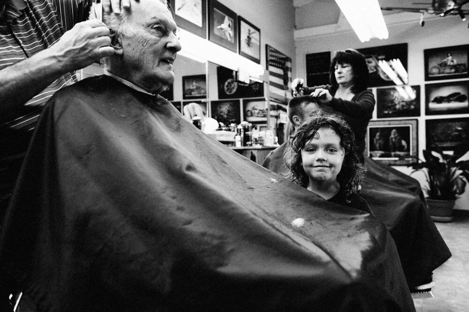 An elderly man receives a haircut in a black and white photo, with a young girl in the foreground and a woman cutting hair in the back.