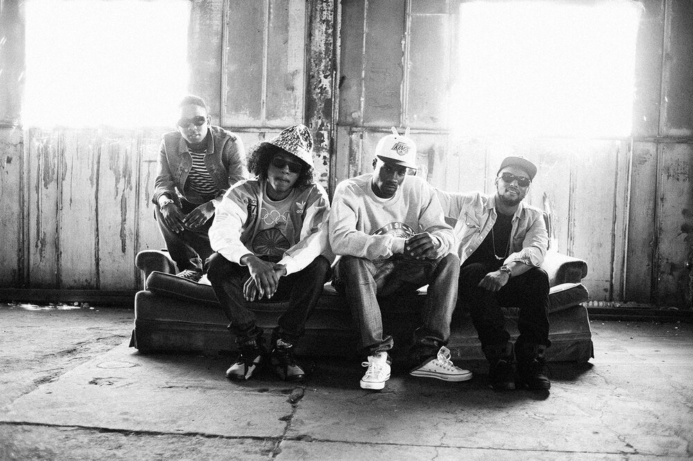 Four men sit together on a worn couch in a bright, industrial-style room, creating a casual yet stylish group portrait in monochrome.