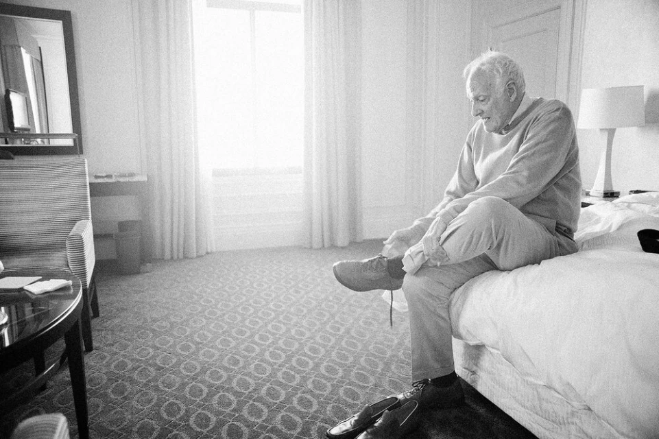 Elderly man sitting on bed, tying shoelaces on casual shoes in naturally lit, monochromatic hotel room with patterned carpet.
