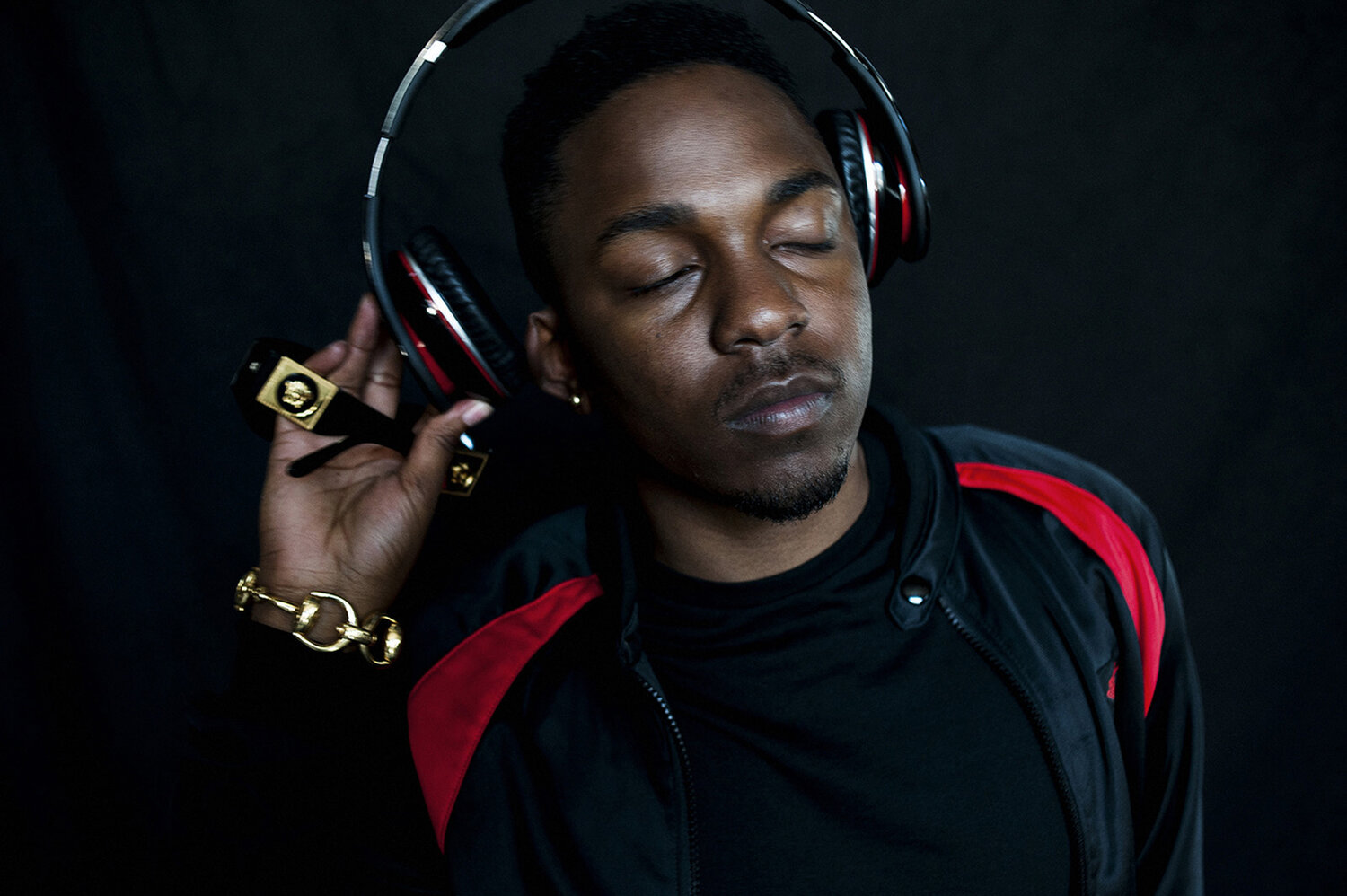 Portrait of a Black male musician with his eyes closed, wearing headphones, sunglasses and a gold chain, against a dark background.