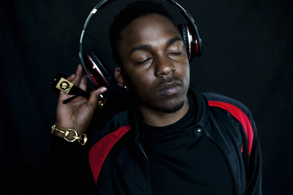 Portrait of a Black male musician with his eyes closed, wearing headphones, sunglasses and a gold chain, against a dark background.