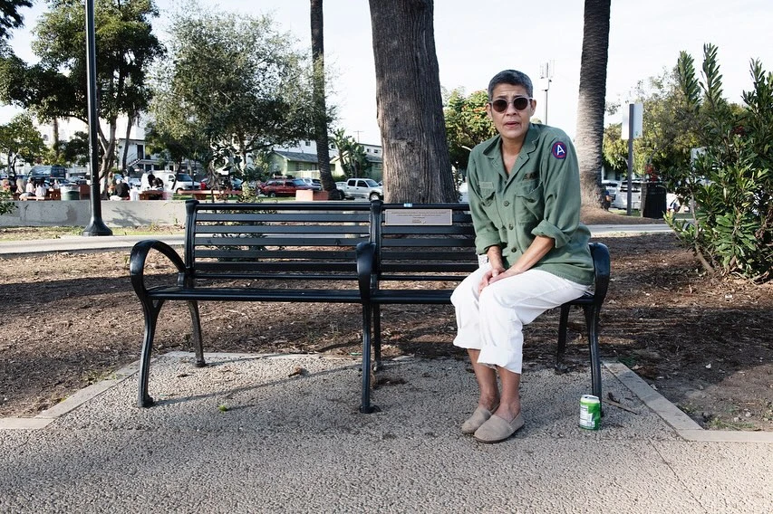 Woman sits on park bench in vintage military jacket, white pants, and sunglasses, with trees and building in the background.