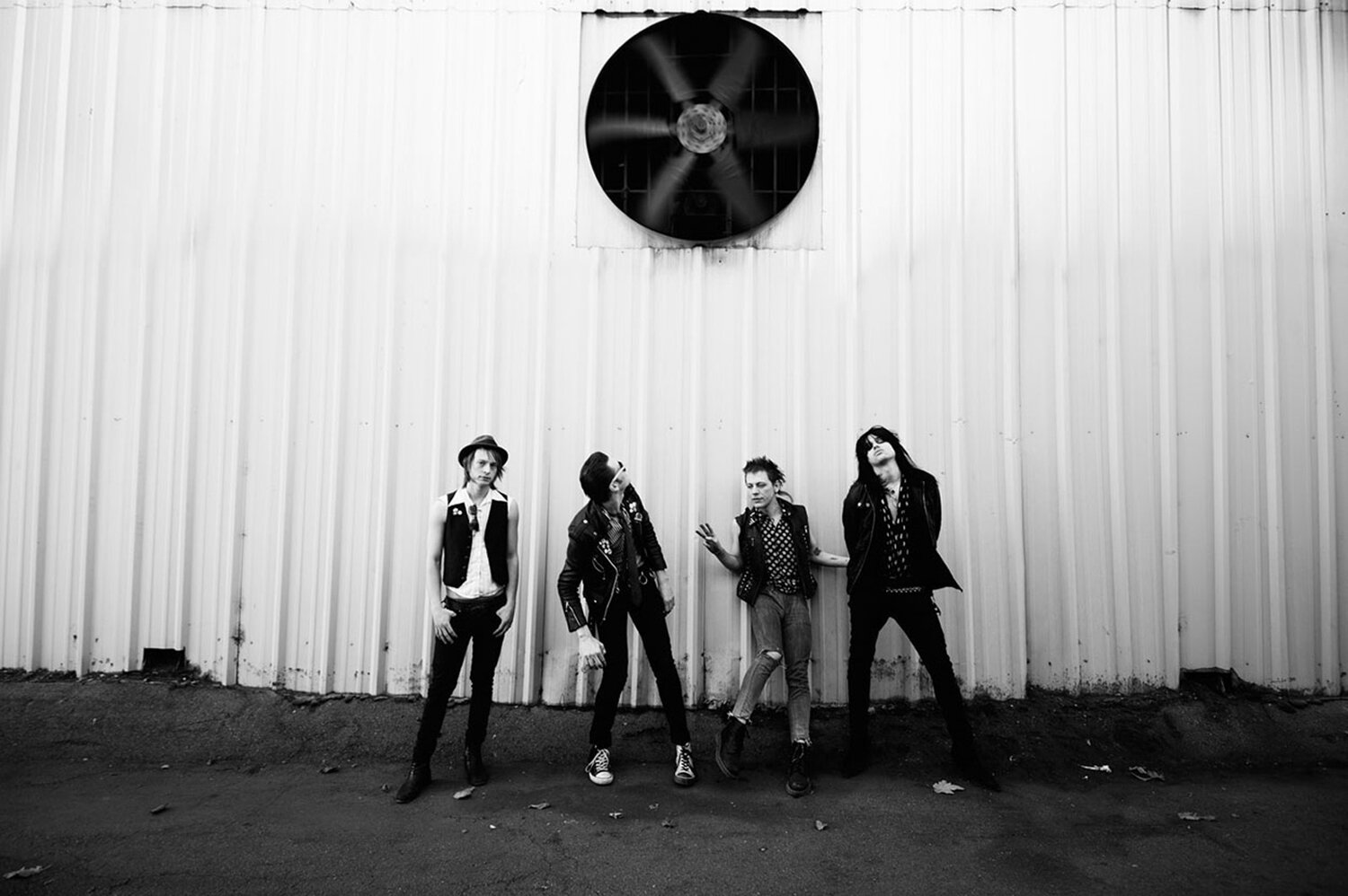 Black and white image of four adult rock musicians posing in black attire against a corrugated metal wall with a large fan.