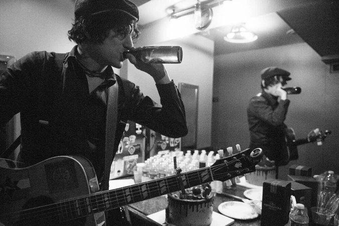 Black and white portrait of a musician playing acoustic guitar and drinking from a bottle, photographed by Jeff Forney