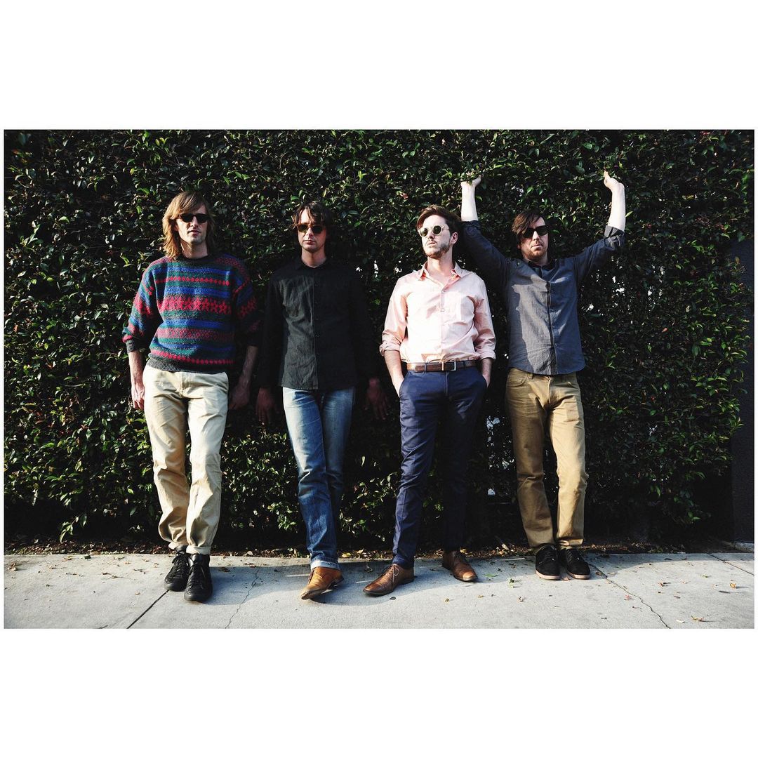 Four male musicians pose outdoors against a green hedge. The group wears casual clothing and sunglasses, with natural lighting.