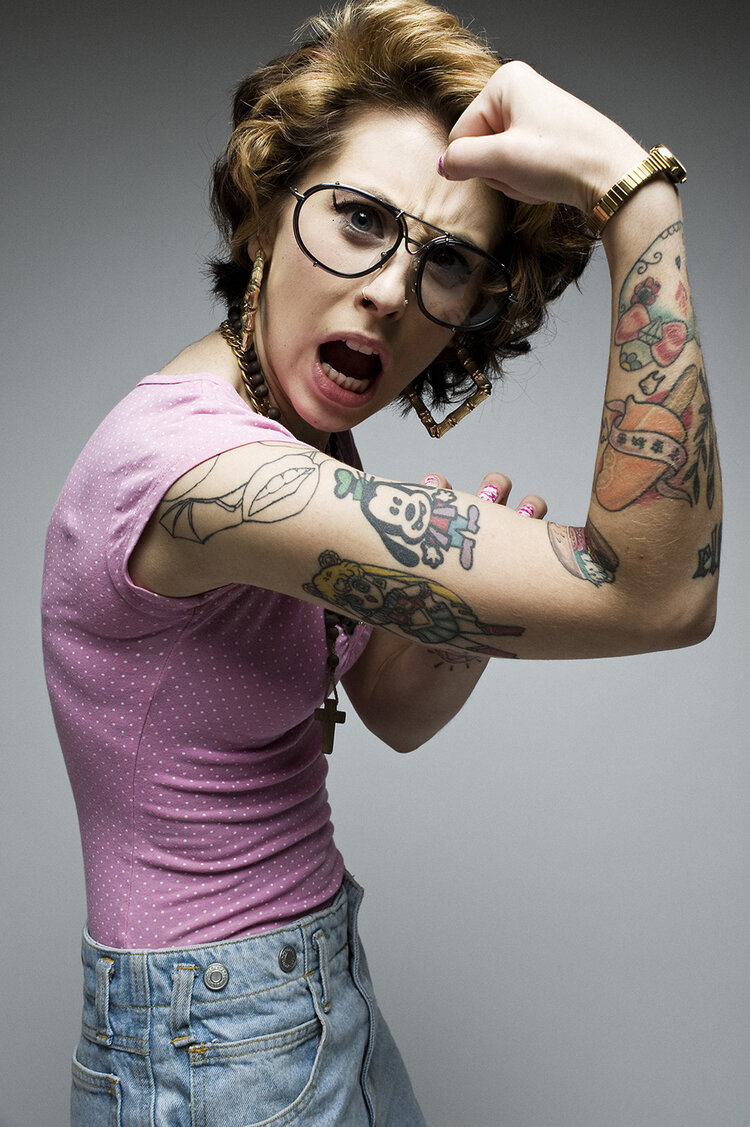 Confident woman with arm tattoos flexes bicep, wearing vintage glasses and gold jewelry against a neutral grey background in studio lighting.