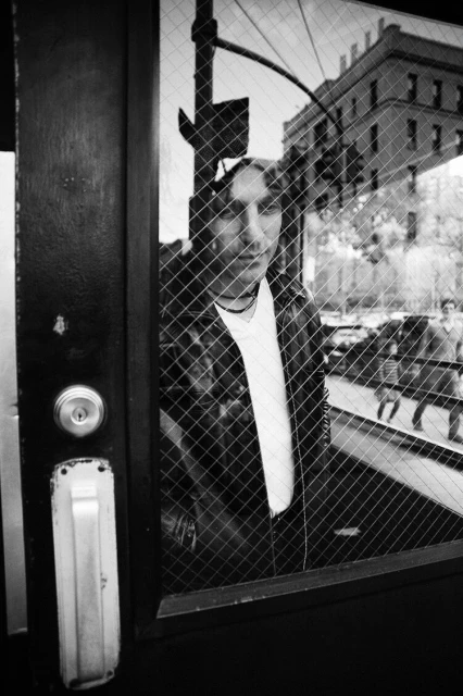 Monochromatic portrait of a man looking out of a window with a metal wire safety screen, city street visible outside.