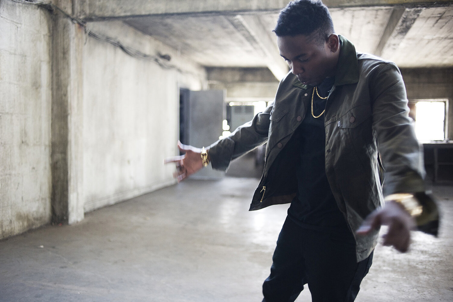 An adult male in a jacket and gold jewelry gestures in an empty concrete room with natural light from windows in an indoor setting.