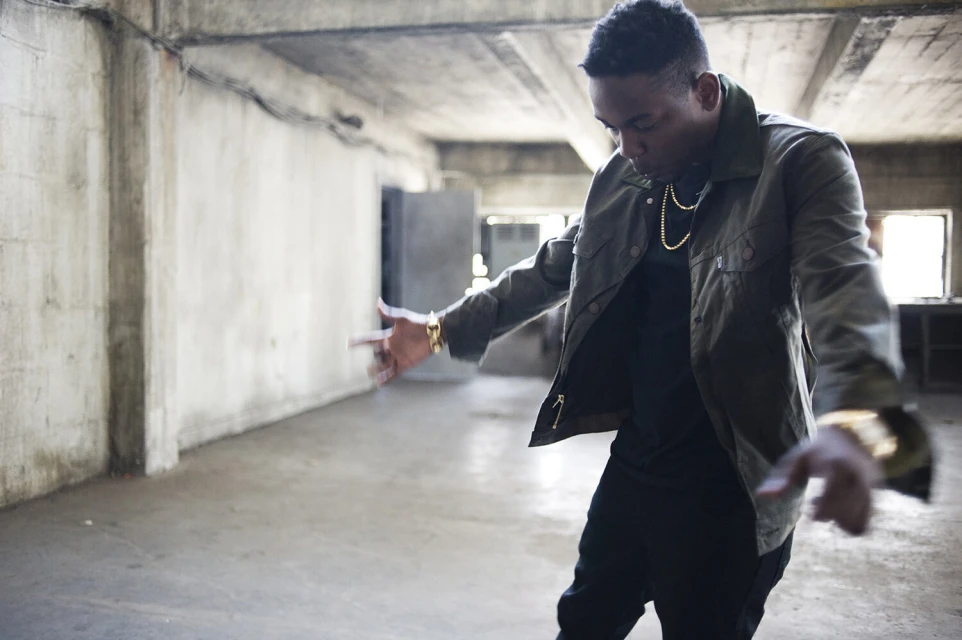 An adult male in a jacket and gold jewelry gestures in an empty concrete room with natural light from windows in an indoor setting.